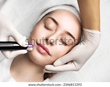 Beautician Doing Permanent Makeup For Lips Of Young Attractive Woman, Beautiful Female Getting Pmu Lip Blushing Treatment In Cosmetologist Cabinet, Professional Artist Using Tattoo Pen Machine Royalty-Free Stock Photo #2346811293