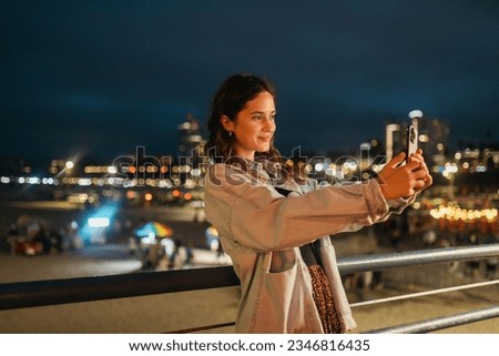 Young beautiful brunette woman with a smartphone making a selfie on an embankment and looking away in the night-illuminated city.