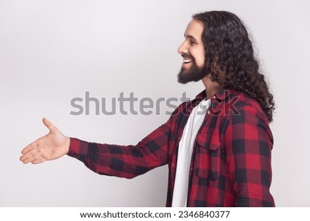 Nice to meet you. Side view portrait of friendly pleased bearded man with long curly hair in checkered red shirt handshaking, smiling happily. Indoor studio shot isolated on gray background.