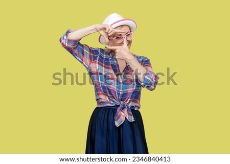 Portrait of positive mature woman wearing checkered shirt, hat and eyeglasses standing with crop composition gesture and looking at target. Indoor studio shot isolated on yellow background.