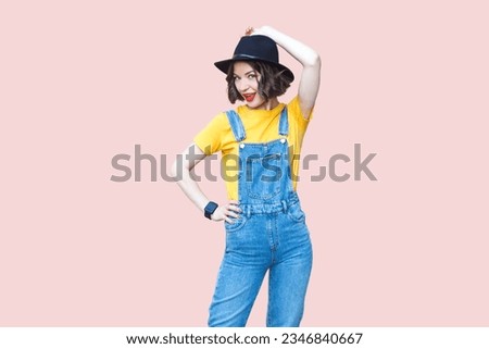 Portrait of funny positive confident hipster woman in blue denim overalls, yellow T-shirt and black hat, smiling to camera. Indoor studio shot isolated on light pink background. Royalty-Free Stock Photo #2346840667