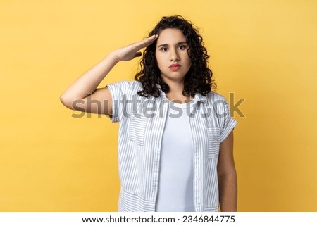 Portrait of responsible serious woman with dark wavy hair saluting commander, listening order with obedient expression, looking at camera. Indoor studio shot isolated on yellow background. Royalty-Free Stock Photo #2346844775