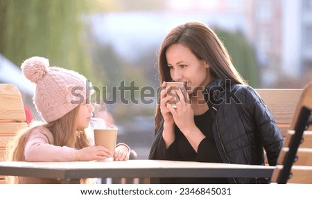 Young pretty woman having good time with her child daughter sitting at street cafe with hot drinks on sunny autumn day. Happiness in family relations concept