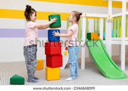 Two girls play with colorful, large, soft blocks-constructor for kindergarten. A fun game and developing skills in building castles and houses. Happy children have fun in the children's room.