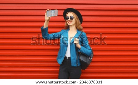 Portrait of stylish smiling young woman taking selfie with mobile phone wearing black round hat, jacket and backpack on red background