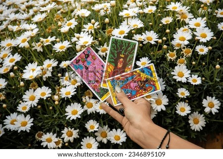 A woman's manicured hand close-up with a chambal bracelet against a background of white daisies holds Tarot cards. 