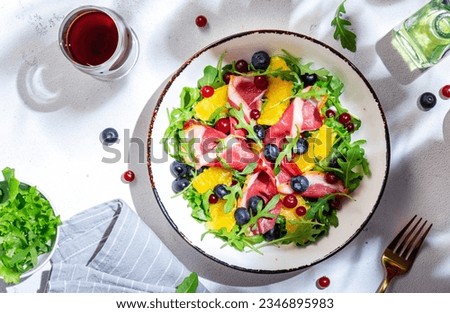 Delicious salad with smoked duck, oranges, blueberries, cranberries and arugula on white table background, top view. Hard sunlight and shadows pattern