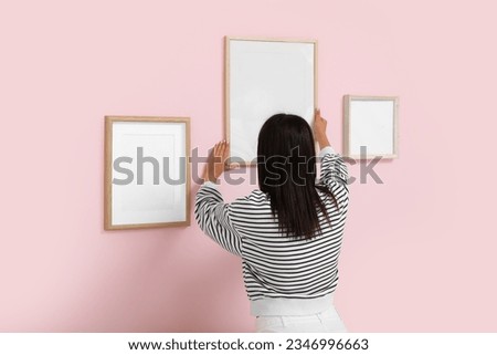Young woman hanging blank frame on pink wall in living room, back view
