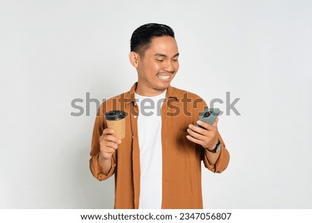 Smiling young Asian man in casual shirt reading news on mobile phone and holding cup of coffee isolated on white background