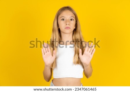 Serious beautiful caucasian kid girl wearing white T-shirt pulls palms towards camera, makes stop gesture, asks to control your emotions and not be nervous