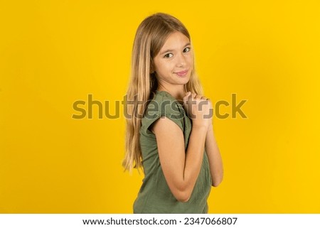 Charming serious beautiful caucasian kid girl wearing green T-shirt keeps hands near face smiles tenderly at camera