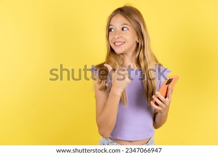 beautiful caucasian kid girl wearing violet T-shirt using and texting with smartphone  pointing and showing with thumb up to the side with happy face smiling