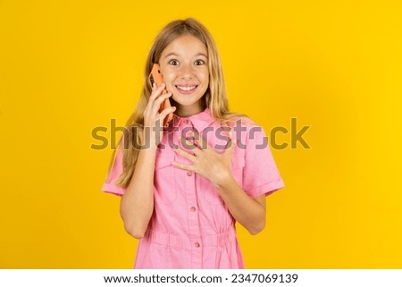 Smiling beautiful caucasian kid girl wearing pink dress talks via cellphone, enjoys pleasant great conversation. People, technology, communication concept