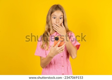 beautiful caucasian kid girl wearing pink dress being deeply surprised, stares at smartphone display, reads shocking news on website, Omg, its horrible! Royalty-Free Stock Photo #2347069153