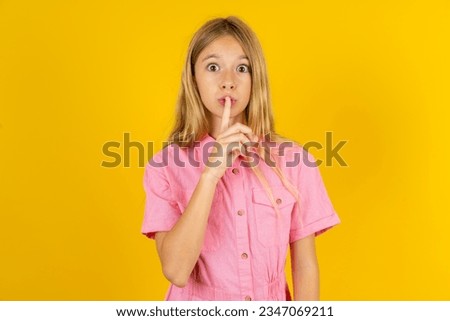 Surprised beautiful caucasian kid girl wearing pink dress makes silence gesture, keeps finger over lips and looks mysterious at camera
