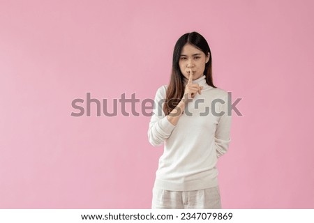 A beautiful Asian woman puts her finger on her lips, showing a shhh, keeping a secret, quiet, whisper, or silence gesture while standing against an isolated pink background. Royalty-Free Stock Photo #2347079869
