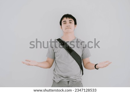 Selective focus shot of young Asian man wearing sage green casual T-Shirt, smartwatch, and sling bag is confuse gesture or don't know with raised arms and flat expression on isolated white background