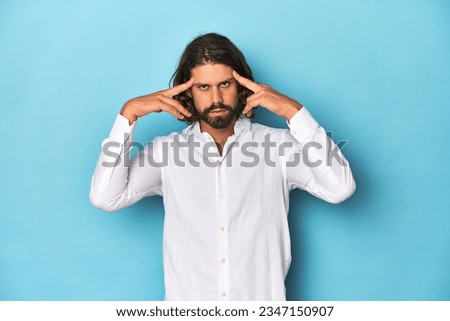 Bearded man in a white shirt, blue backdrop focused on a task, keeping forefingers pointing head.