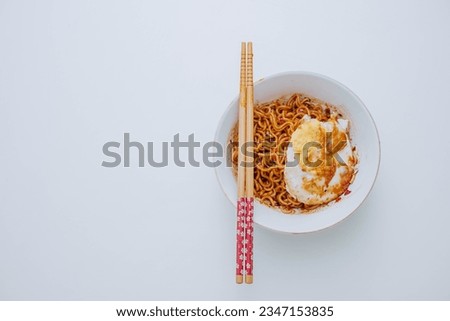 Flat lay or top view shot of a portion of stir-fried Indonesian instant noodles with egg on a white bowl with a pair of wooden chopsticks on white table