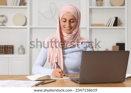 Muslim woman writing notes near laptop at wooden table in room