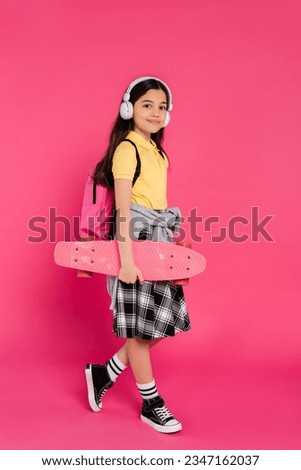 happy schoolgirl in wireless headphones standing with penny board, pink background, after classes