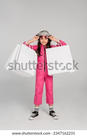 girl in stylish outfit and panama hat holding shopping bags on grey, adjusting pink sunglasses