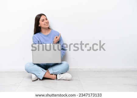 Young caucasian woman with laptop sitting on the floor isolated on white background pointing back