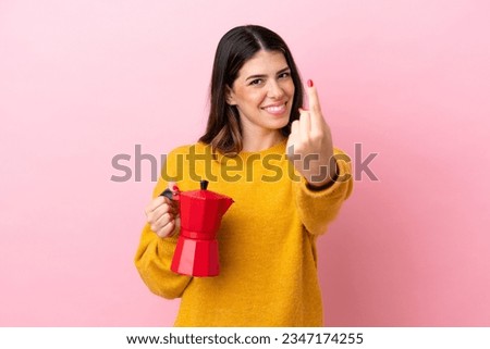 Young Italian woman holding a coffee maker isolated on pink background doing coming gesture