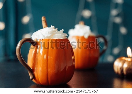 Pumpkin shaped cups of hot drink with whipped cream, cinnamon stick on black table with dark background. Pumpkin latte with spices. Cozy autumn beverage for Thanksgiving, Halloween holidays at home