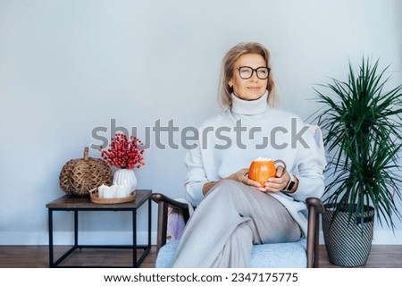 Middle aged woman relaxing with pumpkin shaped cup of hot drink in scandy style hygge interior home with fall mood decor. Lady dreaming, enjoy calm mood without stress, well being alone. Cozy autumn Royalty-Free Stock Photo #2347175775