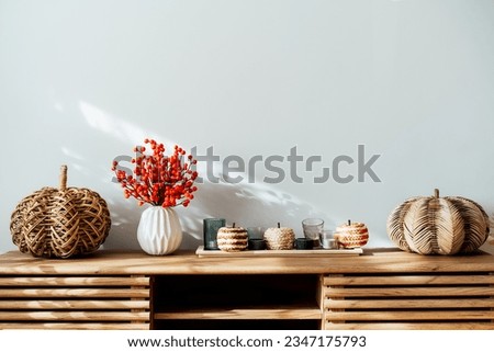 Cozy autumn home interior - various decorative wicker pumpkins, candles, seasonal flowers in vase on the wooden console with white wall background. Scandinavian minimalist hygge home fall decor.