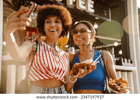 Two beautiful smiling african american women taking selfie while standing outdoors near food court on sunny day