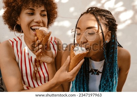 Two cheerful young african american women sharing ice cream while spending time together outdoors on sunny day