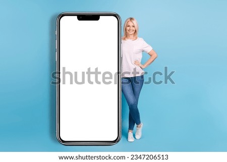 Full size photo of positive pensioner woman white t-shirt jeans sneakers stand near panel empty space isolated on blue color background