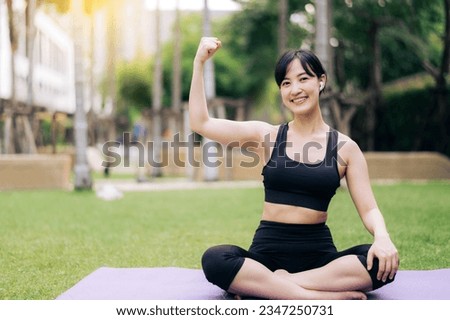 Portrait fit young asian woman wearing black sportswear listening relax music and showing strong health gesture at her arm while do yoga on yoga mat in the outdoor park. Royalty-Free Stock Photo #2347250731