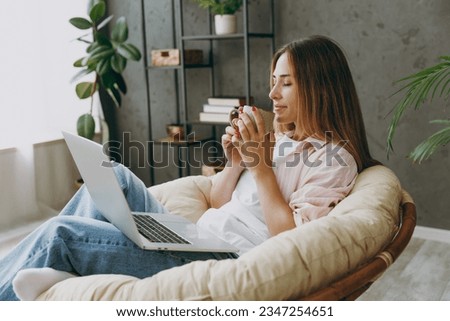 Young IT woman wears casual clothes sits in armchair hold use work on laptop pc computer drink tea stay home hotel flat rest relax spend free spare time in living room indoor. Lifestyle lounge concept