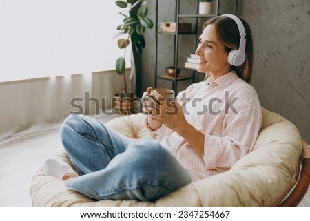 Side view young woman wearing casual clothes sits in armchair listen to music in headphones drink coffee stay at home hotel flat rest relax spend free spare time in living room indoor. Lounge concept Royalty-Free Stock Photo #2347254667