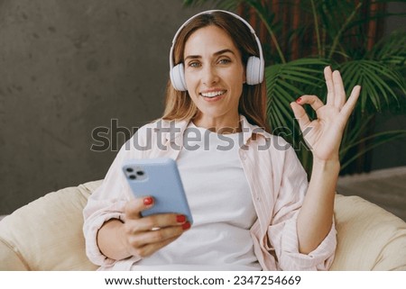Young woman wear casual clothes sit in armchair listen to music in headphones use mobile cell phone show ok okay stay at home hotel flat rest spend free spare time in living room indoor Lounge concept