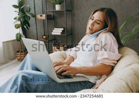 Young IT woman wears casual clothes sits in armchair hold use work on laptop pc computer speak on mobile cell phone stay home hotel flat rest spend free spare time in living room indoor Lounge concept Royalty-Free Stock Photo #2347254681