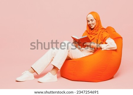 Full body side view happy smiling young arabian asian muslim woman wear orange abaya hijab sit in bag chair read book novel isolated on plain pink background Uae middle eastern islam religious concept