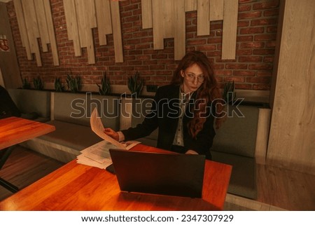 Young lovely redhead woman working on her lap top with one hand and holding a document with the other. Working remotely. Front view