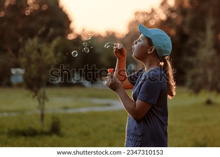 A child of 10-12 years old blows soap bubbles outside. A school-age girl enthusiastically plays in the summer park at sunset.
