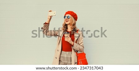 Autumn style outfit, stylish smiling young woman taking selfie with smartphone wearing red french beret hat, jacket, handbag and round sunglasses on gray studio background