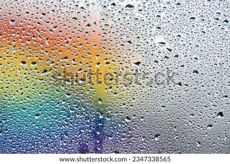 Droplets of rain on a window reveal macro images of the LGBTQ+ Pride Progress flag that is unfocussed in the background. Rainbow colours dominate the scene representing equality for all.