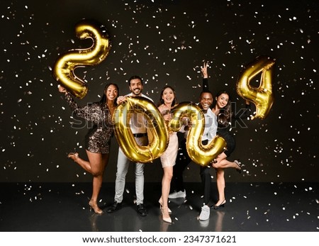 Happy beautiful multiethnic millennials men and woman in nice outfits celebrating New Year 2024 together, holding golden number balloons and posing over black background among confetti