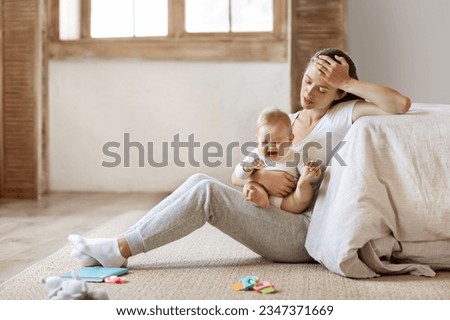 Tired infant baby cries in mother hands, depressed unhappy exhausted mom sitting on floor with crying little child on her lap, bedroom interior, copy space. Postnatal postpartum depression concept Royalty-Free Stock Photo #2347371669