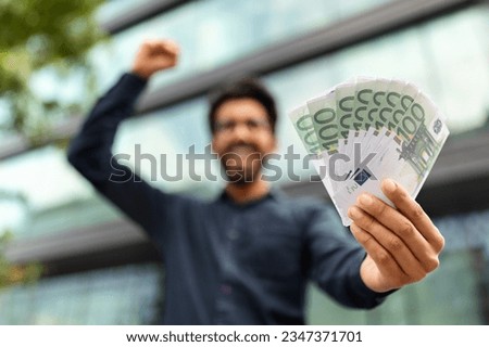 Excited middle eastern businessman holding money cash, recommending investment, trading and cashback, standing outdoors next to office building, selective focus on euro banknotes, copy space Royalty-Free Stock Photo #2347371701