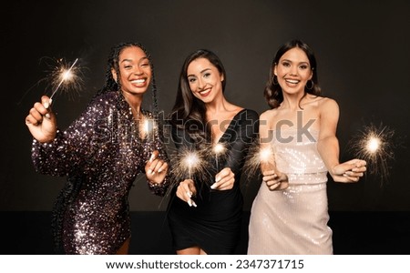 Crazy party time of three beautiful stylish women in elegant outfit celebrating new year 2024 together over black background, young ladies having fun, dancing, holding bengal lights and smiling Royalty-Free Stock Photo #2347371715