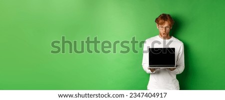 Impressed redhead nerdy guy in glasses showing blank laptop screen and looking at display amazed, standing over green background.