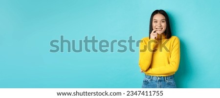 Image of good-looking asian woman in stylish outfit, touching lip and smiling at camera with happy face, standing over blue background.